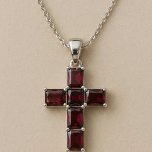 May include: A silver-toned cross pendant necklace. The cross is adorned with rectangular, deep red gemstones. The necklace chain is delicate and also silver-toned. The pendant is centered against a neutral background.