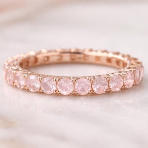 Può includere: Anello eternity in oro rosa con una fila continua di pietre preziose rotonde rosa chiaro. L'anello è posizionato su una superficie di marmo bianco, mettendo in risalto il colore e il design delicati.