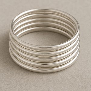 May include: A stack of six silver bangles. The bangles are round and smooth, reflecting light. The jewellery is stacked together, creating a layered effect. The bangles are a simple, elegant accessory.