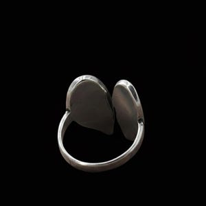 Op de afbeelding: Een zilverkleurige ring met twee ovaalvormige, verhoogde elementen. De ring heeft een gesplitst ontwerp, waarbij de twee ovale vormen naast elkaar zijn geplaatst. De ring staat tegen een zwarte achtergrond, wat de metaalglans benadrukt.