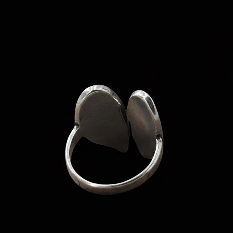 Op de afbeelding: Een zilverkleurige ring met twee ovaalvormige, verhoogde elementen. De ring heeft een gesplitst ontwerp, waarbij de twee ovale vormen naast elkaar zijn geplaatst. De ring staat tegen een zwarte achtergrond, wat de metaalglans benadrukt.