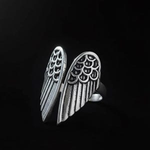 Op de afbeelding: Zilverkleurige ring met een open ontwerp met twee gestileerde vleugelvormen. Elke vleugel heeft gedetailleerde veer- en schubpatronen. De ring staat tegen een donkere achtergrond.