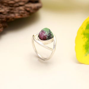 May include: A silver ring featuring a natural ruby-zoisite gemstone. The gemstone is a mix of deep red and green hues, set in a modern, open-design band. The ring is a unique piece of jewelry.