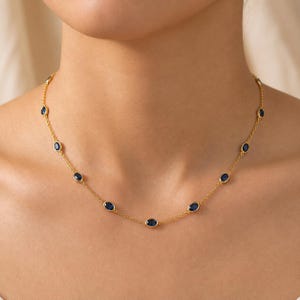 Può includere: Una collana a catena dorata con pietre preziose blu scuro di forma ovale, distanziate uniformemente e incastonate in castoni dorati. La collana è composta da una sottile catena a maglie che sostiene diverse pietre blu disposte simmetricamente.