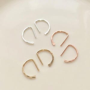 Könnte beinhalten: Drei Paar offene Creolen in Silber-, Gold- und Roségoldtönen. Jeder Ohrring besteht aus einem halbkreisförmigen Drahtdesign mit einem geraden Steg, wodurch eine geometrische Form entsteht. Die Metalloberflächen haben ein glattes, poliertes Finish.