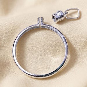 May include: A silver-colored, circular pendant with a small, detached cylindrical clasp and a rectangular loop. The pendant is open in the center and has a smooth, reflective surface. The clasp has a ridged design. The items are on a beige fabric.