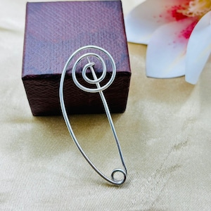 May include: A silver brooch featuring a spiral design and a safety pin clasp. The brooch is set against a dark brown box, with a white and pink flower in the background. This brooch is a unique jewellery accessory.