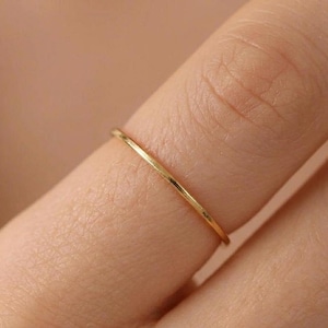 Puede incluir: Un delicado y fino anillo de oro. El anillo es una banda simple, probablemente de oro macizo. Se lleva en un dedo, con el tono de la piel en el fondo. El diseño del anillo es minimalista y elegante.