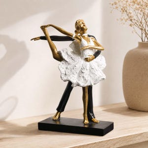 Op de afbeelding: Een decoratief beeldje van twee dansers in een dynamische pose. Eén figuur draagt een getextureerde witte tutu en heeft een goudkleurige huid, terwijl de andere figuur in het zwart is gekleed. Ze staan op een rechthoekige zwarte basis.