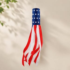 May include: A cylindrical windsock featuring a dark blue top section adorned with white stars. Attached to the base are multiple long, flowing fabric streamers in alternating red and white colors, suspended by a thin white string at the top.