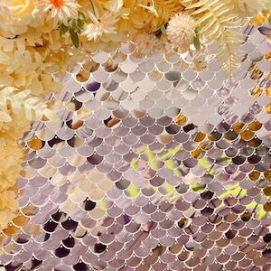 Puede incluir: Primer plano de un telón de fondo decorativo con un patrón brillante en forma de escamas en tonos plateados y dorados. El telón de fondo está adornado con flores artificiales en tonos amarillos y crema suaves, creando un contraste visual atractivo.