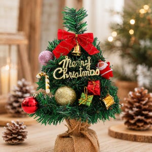 Op de afbeelding: Een kleine kunstmatige groene kerstboom in een met jute omwikkelde voet, versierd met een rode strik, een gouden bel, een gouden Merry Christmas-bordje, een kerstmanfiguur, een zuurstok, een roze glitterbal en miniatuurcadeautjes.