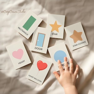 May include: Educational flashcards displaying shapes and objects like a green rectangle, a yellow star, a pink heart, and a red heart balloon. Each card is labelled. A child's hand is partially visible, reaching towards the cards.