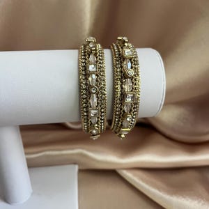 May include: Two gold-toned bangles featuring a textured metal base adorned with rows of small clear crystals and larger square and marquise-shaped clear stones. The bracelets have a detailed, intricate pattern throughout the metal surface.