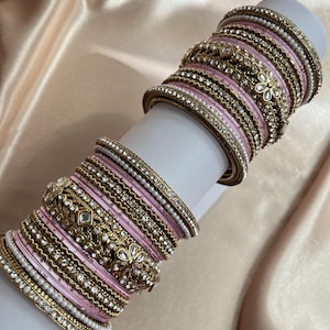May include: A set of stacked bangles featuring alternating bands of light pink satin-like material, gold-toned metal with intricate filigree patterns, and rows of small white faux pearls and clear crystals arranged in floral and geometric designs.