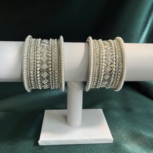 May include: Two sets of silver-toned bangles featuring intricate metalwork, rows of small white faux pearls, and embedded clear crystals arranged in a geometric diamond pattern. Each set consists of multiple stacked bands with textured detailing.