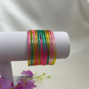May include: A set of eleven thin, translucent bangles in vibrant shades of pink, green, and yellow. The circular bracelets feature a smooth, glossy finish and are arranged in a stacked formation, showcasing their bright, semi-transparent colours.