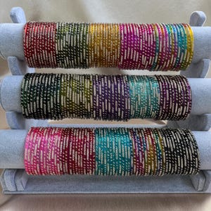 May include: A collection of thin metal bangles arranged in three horizontal rows on a display stand. Each bangle features a vibrant, solid colour finish with a repeating pattern of small, silver-toned dots encircling the entire circumference.