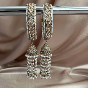 May include: A pair of gold-toned bangles featuring a repeating floral pattern set with clear stones and bordered by small white beads. Each bangle is attached to a bell-shaped jhumka pendant adorned with clear stones and long white bead tassels.