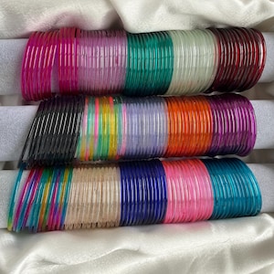 May include: A collection of thin, translucent glass bangles arranged in three horizontal rows. The bangles feature a wide spectrum of vibrant colours including pink, purple, green, white, red, black, yellow, orange, blue, and clear tones.