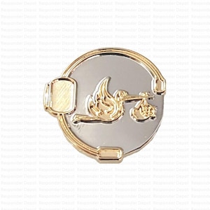 May include: A silver and gold-toned metal pin featuring a stork carrying a baby in a bundle. The pin has a circular design with a rectangular accent on the left side. The stork and baby are in gold, set against a silver background.