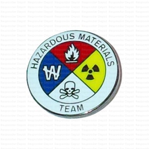 May include: A round, silver-colored Hazardous Materials Team badge. The badge is divided into four quadrants with symbols for fire, radiation, and poison. The text "HAZARDOUS MATERIALS" and "TEAM" are displayed.