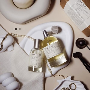 May include: Two bottles of Santal 33 perfume, one large and one small, are displayed in an overhead shot. A white seashell-shaped dish holds gold jewellery. A lit candle sits in a white holder. A small box and a pearl necklace are also present.