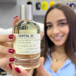 May include: A clear glass bottle of Santal 33 eau de parfum with a silver cap. The label includes the text "SANTAL 33" and "100ml 100 ml". The bottle is held in front of a person.