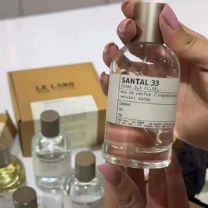 May include: A clear glass bottle of Santal 33 eau de parfum, 100ml (3.4 fl oz), with a silver cap. The label reads "natural spray." Several other perfume bottles are visible in the background, along with a Le Labo box. The bottle is held by a person.