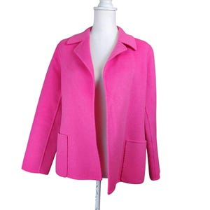 May include: A bright hot pink open-front jacket featuring a wide notched collar, long sleeves, and two large rectangular patch pockets on the front. The garment is displayed on a white mannequin, showcasing a solid color and a textured fabric finish.