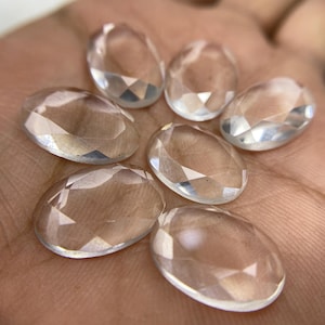 Pode incluir: Sete pedras preciosas ovais transparentes e facetadas são dispostas em um padrão circular. As pedras são transparentes com múltiplas facetas que capturam a luz. As pedras são exibidas em um fundo cor de pele.