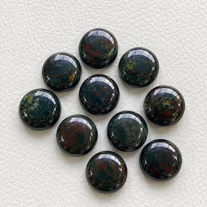 Puede incluir: Una colección de diez piedras preciosas redondas y pulidas. Las piedras son de un verde intenso con inclusiones rojas y marrones, y tienen una superficie brillante y reflectante. Las piedras están dispuestas en un patrón ligeramente disperso.