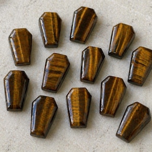 Può includere: Una collezione di ciondoli a forma di bara in pietra occhio di tigre. I ciondoli sono un mix di marrone e oro con finitura lucida. Le pietre sono disposte su una superficie chiara.