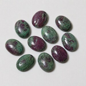 May include: A collection of ten oval-shaped gemstones. The stones have a polished surface and a mix of deep burgundy and green colors. The gemstones are arranged on a white surface, showcasing their natural beauty and unique patterns.