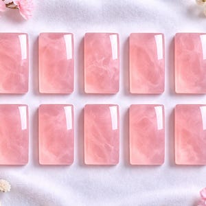 Può includere: Dieci pietre rettangolari di quarzo rosa con finitura lucida. Le pietre rosa sono disposte su una superficie bianca, con fiori rosa e fiori bianchi agli angoli.