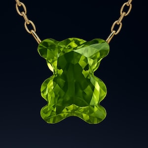 Peut inclure: Un collier en chaîne dorée avec un pendentif en pierre précieuse vert lime facetté en forme d'ours en peluche. Le pendentif présente un design géométrique étincelant. Le fond est bleu marine foncé.