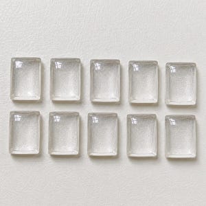 Puede incluir: Diez baldosas de vidrio rectangulares transparentes, dispuestas en dos filas de cinco. Cada baldosa tiene esquinas redondeadas y una superficie ligeramente texturizada, que refleja la luz. Sobre un fondo neutro blanquecino.