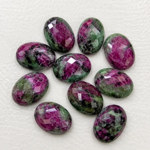 May include: A collection of nine oval-shaped, faceted ruby zoisite gemstones. Each stone displays a distinct mottled pattern of vibrant magenta-pink ruby inclusions contrasted against deep forest green zoisite and dark black mineral flecks.