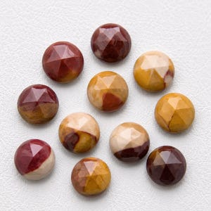 May include: A collection of eleven round, faceted cabochons made of natural stone. Each piece features a unique blend of earthy colors, including shades of deep maroon, mustard yellow, cream, and brown, with a polished, reflective surface finish.