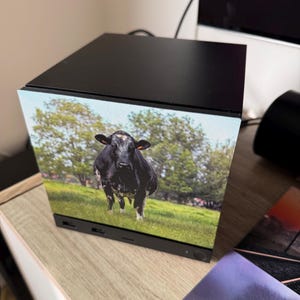 May include: A black cube-shaped computer case featuring a high-resolution printed image of a black and white cow standing in a green pasture with trees in the background. The front panel displays the cow, while the top and base are solid black.