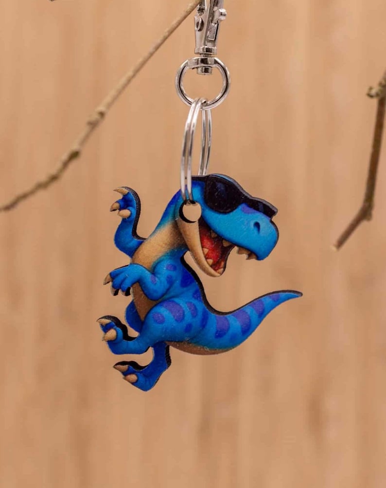May include: A blue dinosaur keyring featuring a T-Rex character with dark blue spots, a tan underbelly, and black sunglasses. The dinosaur has an open mouth showing teeth and is attached to a silver metal split ring and a lobster claw clasp.