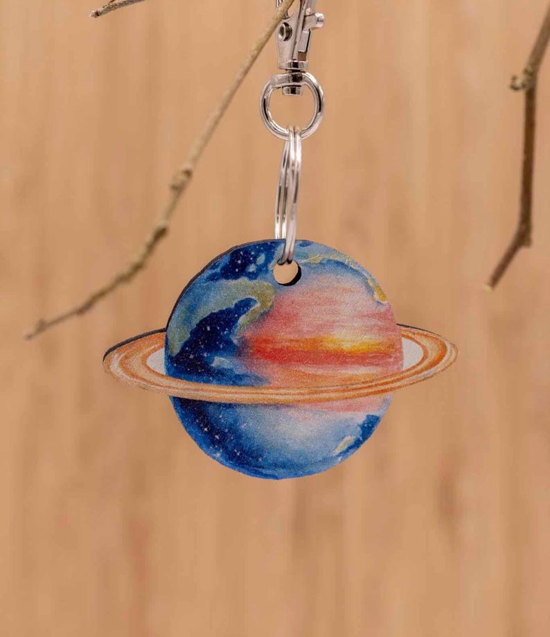 K&ouml;nnte beinhalten: Ein runder Schl&uuml;sselanh&auml;nger mit einer farbenfrohen Illustration eines Planeten mit Ringen. Die Planetenoberfl&auml;che zeigt Schattierungen von tiefem Blau, Wei&szlig; und einen leuchtenden orange-roten Farbverlauf. Er ist an einem silberfarbenen Metallring befestigt.
