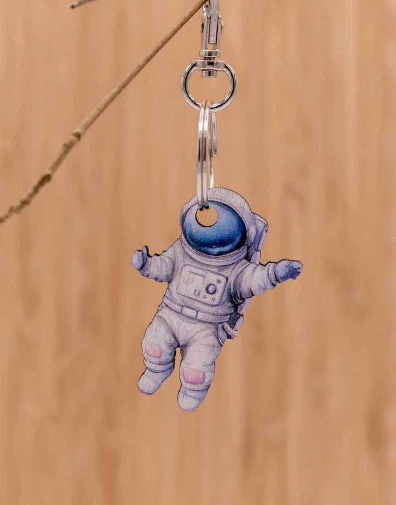 Puede incluir: Un llavero con una figura de astronauta plana e ilustrada, con un traje blanco y una visera de casco azul oscuro. El astronauta est&aacute; suspendido de una anilla de metal plateado y un cierre de mosquet&oacute;n, mostrando un dise&ntilde;o detallado.