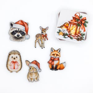 May include: A set of five wooden animal cutouts including a raccoon in a Santa hat, a fawn with a red bow, a hedgehog holding a gift, a rabbit in a Santa hat, and a fox in a scarf, alongside a square tin featuring a lantern and robin.