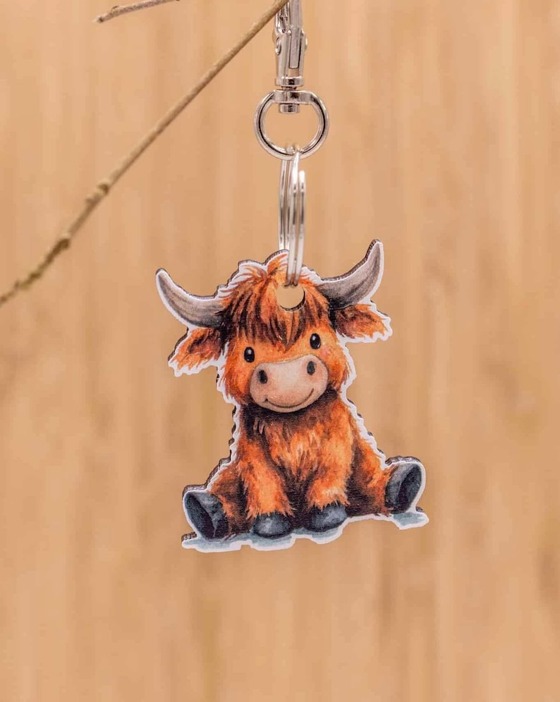 May include: A keychain featuring a flat, die-cut illustration of a fluffy, orange Highland cow sitting down. The cow has long, curved horns, dark eyes, and black hooves. It is attached to a silver-toned metal split ring and a swivel lobster clasp.