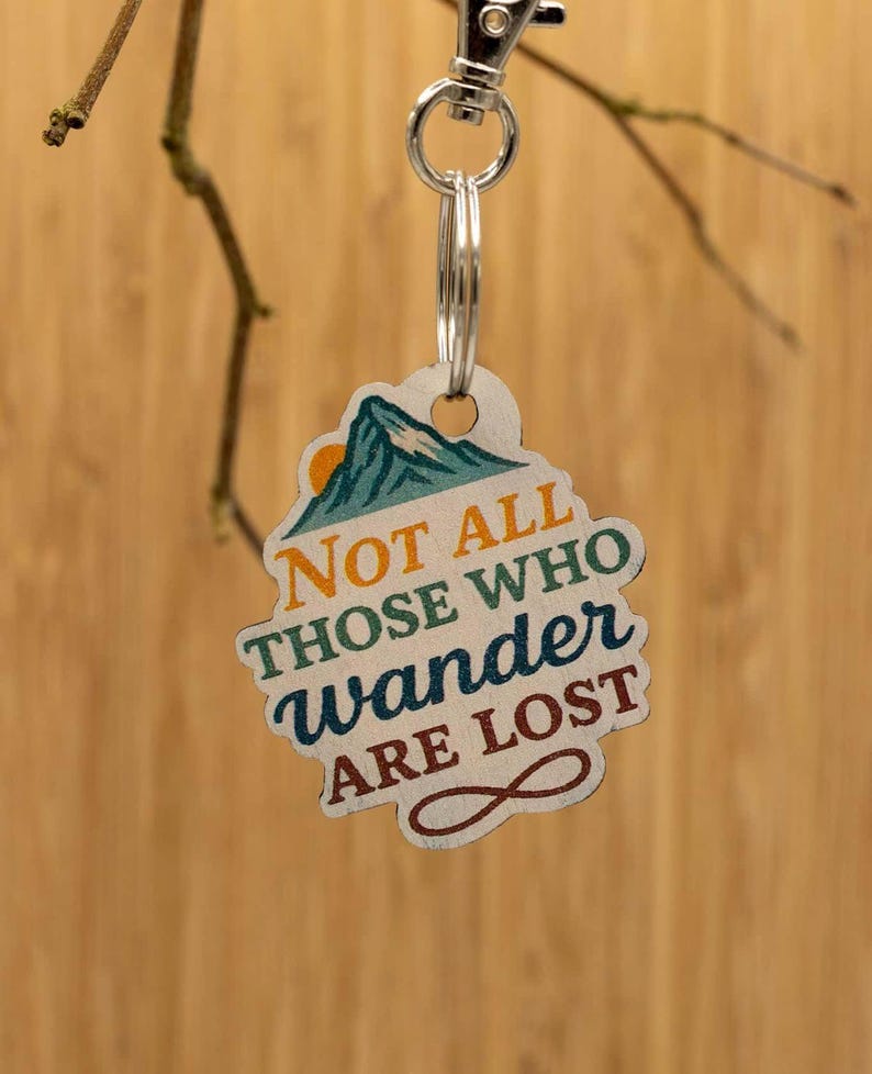 Puede incluir: Un llavero de madera con un gr&aacute;fico de monta&ntilde;a, un peque&ntilde;o sol naranja y el texto "NOT ALL THOSE WHO wander ARE LOST" en letras multicolores, acabado con un mosquet&oacute;n de metal plateado y una anilla circular para llaves.
