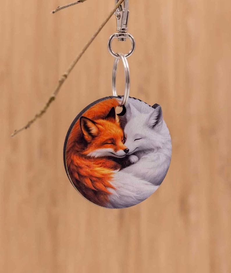 May include: A circular keychain featuring a printed illustration of an orange fox and a white fox curled together in a sleeping position. The keychain is attached to a silver-colored metal split ring and a lobster clasp hardware mechanism.