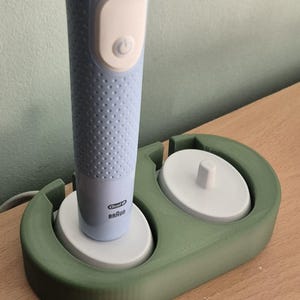Peut inclure: Une brosse à dents électrique Oral-B Braun bleu clair dans un support vert. Le support comporte deux espaces circulaires, un pour la brosse à dents et un pour une tête de rechange. La brosse à dents a un bouton d'alimentation et le nom de la marque.