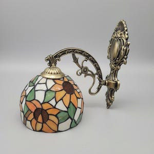 May include: A decorative wall sconce with a stained glass lampshade featuring a sunflower design in orange, yellow, green, and white. The metal fixture has an ornate, antique bronze finish. The lamp is designed to be mounted on a wall.