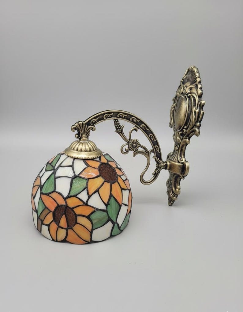 May include: A decorative wall sconce with a stained glass lampshade featuring a sunflower design in orange, yellow, green, and white. The metal fixture has an ornate, antique bronze finish. The lamp is designed to be mounted on a wall.